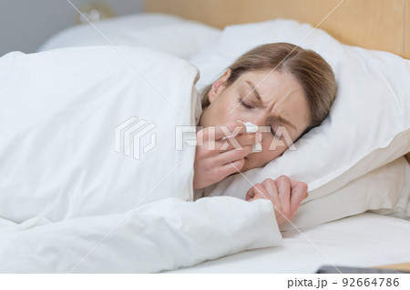 Close-up photo of a sick woman lying under a blanket in bed at home, has a runny nose and cold Close-up photo of a sick woman lying under a blanket in bed at home, has a runny nose and cold 92664786