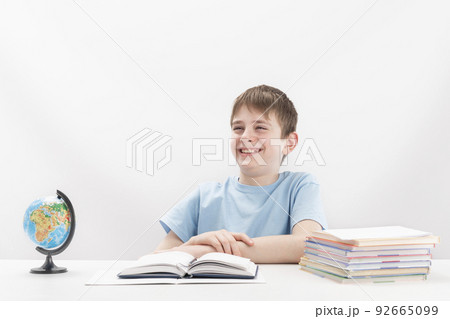 Cheerful laughing teenage boy sits at table while doing his homework. Stack of notebooks and globe on table. International education. Cheerful laughing teenage boy sits at table while doing his homework. Stack of notebooks and globe on table. International education. 92665099