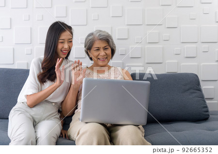 Young asian woman and her grandmother waving hand via remote communication through laptop computer in the living room. Happy family living together 92665352