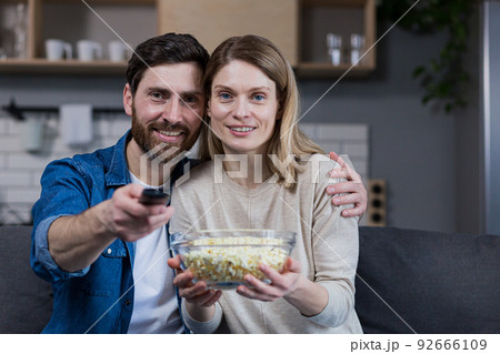 Photo of close couple man and woman together at home having fun and happily watching TV and eating popcorn Photo of close couple man and woman together at home having fun and happily watching TV and eating popcorn 92666109