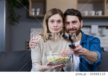 Photo of close couple man and woman together at home having fun and happily watching TV and eating popcorn 92666110