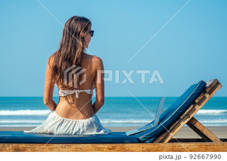 beautiful and sexy young woman sitting in lotus pose and medetiruet on a lounger with a laptop by the sea. woman freelancer doing yoga with a laptop in the resort beautiful and sexy young woman sitting in lotus pose and medetiruet on a lounger with a laptop by the sea. woman freelancer doing yoga with a laptop in the resort 92667990