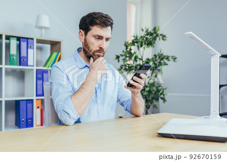 Upset and sad businessman looking at phone screen, man working in bright office Upset and sad businessman looking at phone screen, man working in bright office 92670159