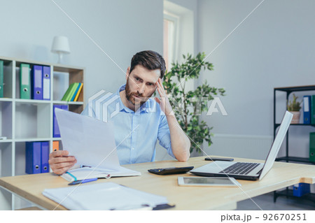 Pensive businessman freelancer at home, works at paperwork, examines thinking documents, husband works at home office at desk, uses laptop Pensive businessman freelancer at home, works at paperwork, examines thinking documents, husband works at home office at desk, uses laptop 92670251