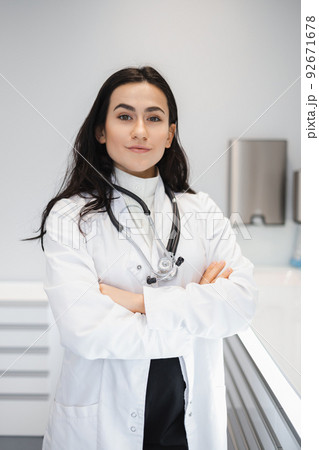 Portrait of female doctor in lab coat standing with crossed hands on her office Portrait of female doctor in lab coat standing with crossed hands on her office 92671678