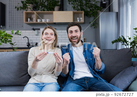 Happy married couple man and woman looking at the camera and rejoicing, holding a positive pregnancy test, celebrating expecting a baby 92672727
