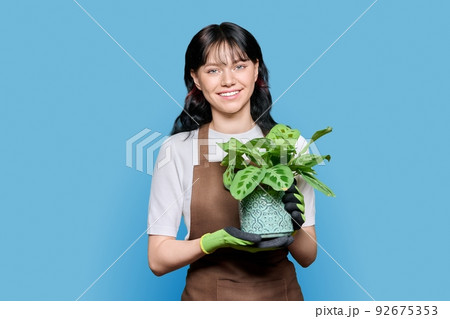 Smiling young woman in apron gloves with potted plant on blue isolated background 92675353