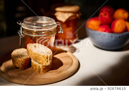 Still life. Slices of whole grain bread on a wooden board, a jar with homemade jam and ripe apricots on a white table 92676610