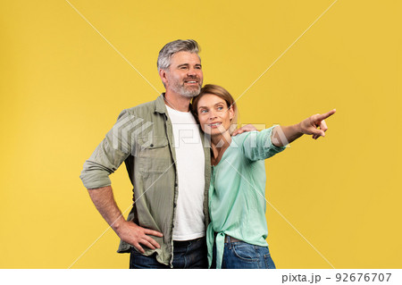 Look there. Happy mature spouses embracing and woman pointing with finger at copy space, yellow background Look there. Happy mature spouses embracing and woman pointing with finger at copy space, yellow background 92676707