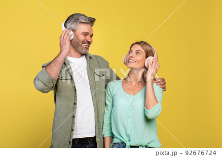 Happy middle aged couple in headphones listening to music, hugging and looking at each other over yellow background Happy middle aged couple in headphones listening to music, hugging and looking at each other over yellow background 92676724
