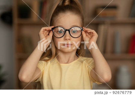 Portrait Of Little Girl Wearing Eyeglasses Sitting In Library Portrait Of Little Girl Wearing Eyeglasses Sitting In Library 92676779