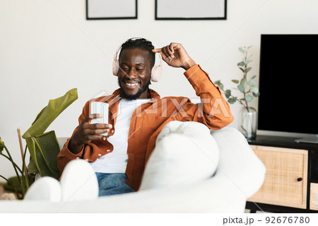 Excited black guy in headphones enjoying music on smartphone and scrolling social networks feed, resting on sofa at home Excited black guy in headphones enjoying music on smartphone and scrolling social networks feed, resting on sofa at home 92676780
