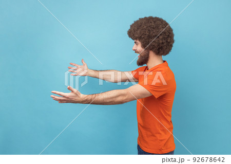 Come into my arms. Side view of satisfied man with Afro hairstyle wearing orange T-shirt reaching out to camera, stretching arms to hug you. Indoor studio shot isolated on blue background. 92678642