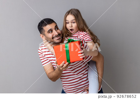 Portrait of pleased father holding his surprised daughter on his back and giving her a present for birthday, family wearing striped T-shirts. Indoor studio shot isolated on gray background. 92678652