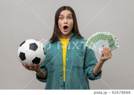 Shocked amazed woman showing soccer ball and fun of hundred euro bills, winning lot of money betting for sport, wearing casual style jacket. Indoor studio shot isolated on gray background. 92678666