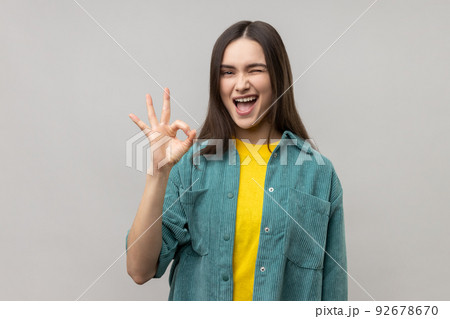 Portrait of satisfied beautiful young woman standing, looking at camera showing Ok sign gesture and winking, wearing casual style jacket. Indoor studio shot isolated on gray background. 92678670
