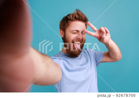 Delighted positive optimistic bearded man taking selfie, looking at camera POV, point of view of photo, showing victory gesture and tongue out. Indoor studio shot isolated on blue background. Delighted positive optimistic bearded man taking selfie, looking at camera POV, point of view of photo, showing victory gesture and tongue out. Indoor studio shot isolated on blue background. 92678700