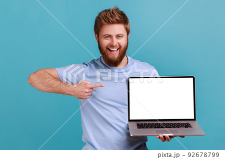 Portrait of happy bearded man holding laptop and pointing at empty screen, smiling satisfied with computer app, looking at camera. Indoor studio shot isolated on blue background. 92678799