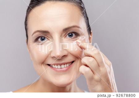 Closeup portrait of middle aged woman applies face cream, enjoys new anti wrinkle cosmetic product, prevents sign of skin aging, wears minimal makeup. Indoor studio shot isolated on gray background. 92678800