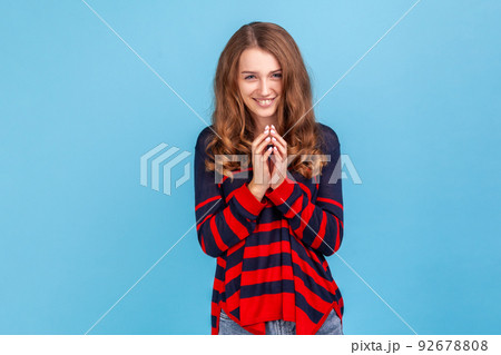 Portrait of young devious woman wearing striped casual style sweater looking at camera with cunning tricky face and smirk, planning evil trick. Indoor studio shot isolated on blue background. 92678808