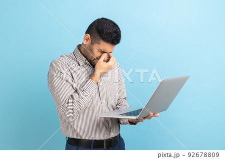 Portrait of young adult businessman standing with laptop in hands, rubbing his eyes, looks tired and exhausted, wearing striped shirt. Indoor studio shot isolated on blue background. 92678809