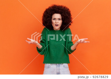What do you want. Woman with Afro hairstyle wearing green casual sweater asking who why make this conflict, looking with annoyed indignant expression. Indoor studio shot isolated on orange background. 92678829