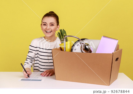 Happy woman office worker sitting at workplace with cardboard box with her things and smiling, having new job, signs an agreement. Indoor studio studio shot isolated on yellow background. 92678844