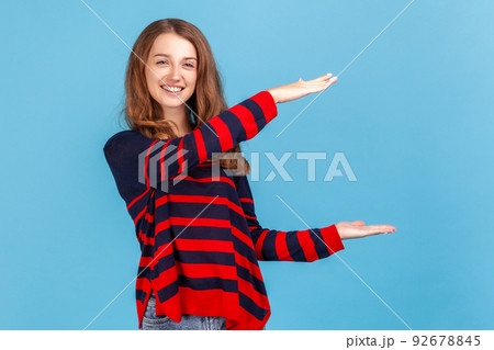 Optimistic woman wearing striped casual style sweater, presenting copy space between hands, holding empty place for commercial text or image. Indoor studio shot isolated on blue background. 92678845