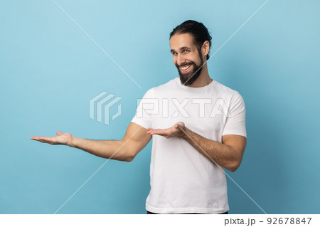Portrait of satisfied optimistic handsome man with beard in white T-shirt standing with wide raised arms and welcoming or sharing. Indoor studio shot isolated on blue background. 92678847