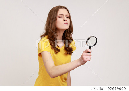 Young attractive curious female with brown hair looking through magnifying glass, spying, finding out something, inspecting crime scene. Indoor studio shot isolated on gray background. 92679000