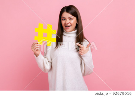 Female points at yellow hashtag symbol, promoting viral topic in social network, tagging blog trends, wearing white casual style sweater. Indoor studio shot isolated on pink background. Female points at yellow hashtag symbol, promoting viral topic in social network, tagging blog trends, wearing white casual style sweater. Indoor studio shot isolated on pink background. 92679016