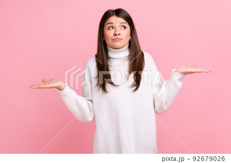Ambiguous confused dark haired female standing with raised arms, showing helpless gesture, no answer, wearing white casual style sweater. Indoor studio shot isolated on pink background. Ambiguous confused dark haired female standing with raised arms, showing helpless gesture, no answer, wearing white casual style sweater. Indoor studio shot isolated on pink background. 92679026