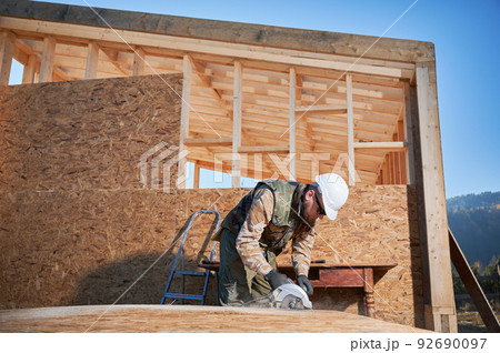 Carpenter using circular saw for cutting wooden OSB board. Man worker building wooden frame house. Carpentry concept. Carpenter using circular saw for cutting wooden OSB board. Man worker building wooden frame house. Carpentry concept. 92690097