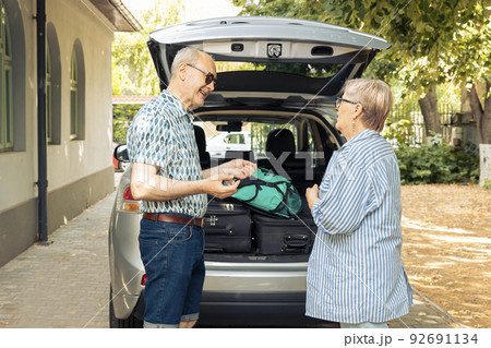 Old husband and wife sitting in driveway with car, leaving on holiday vacation adventure after loading travelling bags and suitcases in automobile trunk. Leaving on journey with trolley for leisure. 92691134