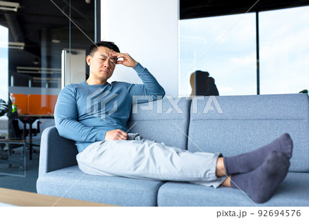 Tired and pensive Asian worker, resting on the office sofa, in a modern office, a businessman makes a difficult and fateful decision Tired and pensive Asian worker, resting on the office sofa, in a modern office, a businessman makes a difficult and fateful decision 92694576