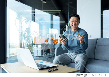 Happy asian male businessman holding credit card and phone, online shopping worker sitting in office 92694594