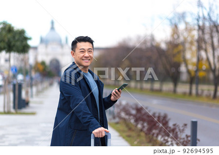 Happy Asian tourist, warmly dressed, in a European city, looks at the camera and smiles, uses the application on the phone to navigate the city 92694595