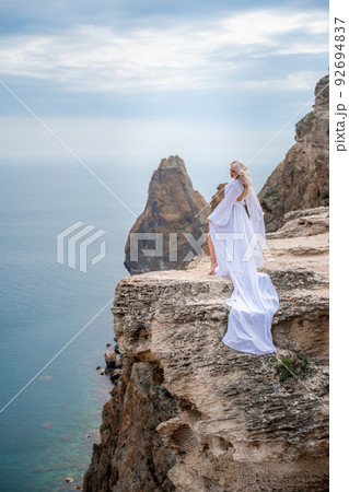 Blonde with long hair on a sunny seashore in a white flowing dress, rear view, silk fabric waving in the wind. Against the backdrop of the blue sky and mountains on the seashore. Blonde with long hair on a sunny seashore in a white flowing dress, rear view, silk fabric waving in the wind. Against the backdrop of the blue sky and mountains on the seashore. 92694837