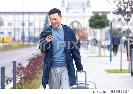 Asian tourist, male passenger arrives from the airport, walks around the city with a suitcase, a man smiles and rejoices uses the phone and the application to book accommodation and book a taxi Asian tourist, male passenger arrives from the airport, walks around the city with a suitcase, a man smiles and rejoices uses the phone and the application to book accommodation and book a taxi 92695184