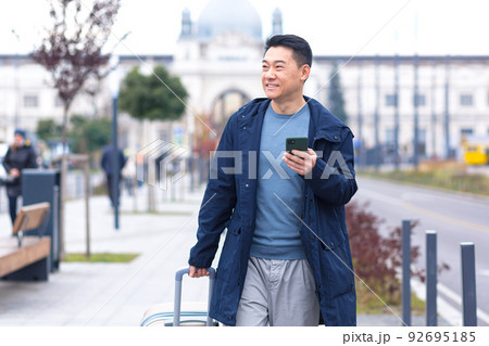 Asian tourist, male passenger arrives from the airport, walks around the city with a suitcase, a man smiles and rejoices uses the phone and the application to book accommodation and book a taxi Asian tourist, male passenger arrives from the airport, walks around the city with a suitcase, a man smiles and rejoices uses the phone and the application to book accommodation and book a taxi 92695185