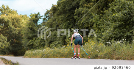 Web banner of biathlon. Athletic young woman training on the roller ski with poles and helmet at country road, back view. Low angle view. Copy space. Concept of sports competition and summer workout Web banner of biathlon. Athletic young woman training on the roller ski with poles and helmet at country road, back view. Low angle view. Copy space. Concept of sports competition and summer workout 92695460