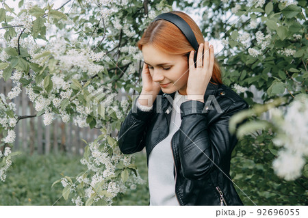 A red-haired teenage girl listens to music in headphones, configured via the phone. The time of spring flowering of cherry orchards A red-haired teenage girl listens to music in headphones, configured via the phone. The time of spring flowering of cherry orchards 92696055