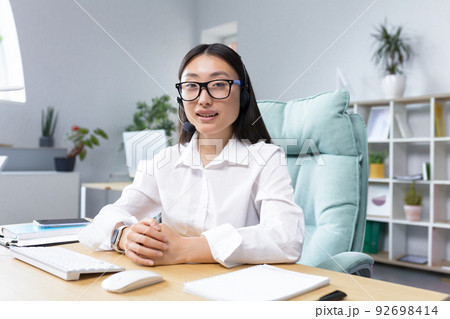 Portrait of a young beautiful Asian woman in glasses and headphones with a microphone, sitting in the office at the desk conducting an online webinar, consulting, telling, gesturing with her hands. Portrait of a young beautiful Asian woman in glasses and headphones with a microphone, sitting in the office at the desk conducting an online webinar, consulting, telling, gesturing with her hands. 92698414