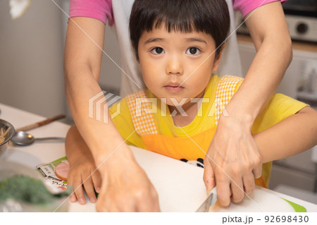 お母さんに、子供用包丁できゅうりを切る手解きを受けている最中によそ見する4歳男児 92698430