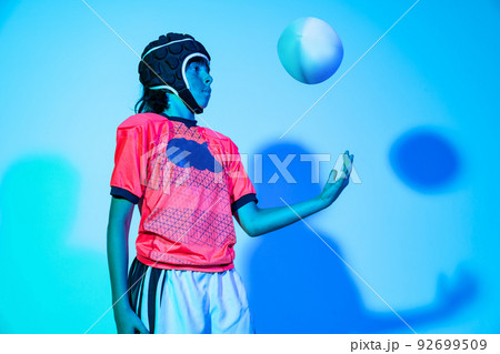 Portrait of junior male rugby player in sports uniform psoing with ball isolated on blue background in neon light. Sport, art, creativity concept Portrait of junior male rugby player in sports uniform psoing with ball isolated on blue background in neon light. Sport, art, creativity concept 92699509
