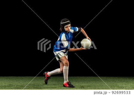 Dynamic portrait of school age boy, junior male rugby player practicing rugby football isolated on dark background with grass floooring. Dynamic portrait of school age boy, junior male rugby player practicing rugby football isolated on dark background with grass floooring. 92699533