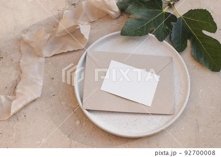 Summer branding, wedding stationery. Mediterranean design. Business card, invitation mock up on craft envelope and ceramic plate. Green fig leaves and silk ribbon. Beige marble background. Flatlay 92700008