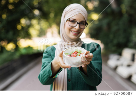 Focus on bowl with salad. Young charming muslim woman holding a c bowl with fresh organic salad Focus on bowl with salad. Young charming muslim woman holding a c bowl with fresh organic salad 92700087