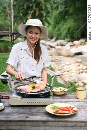 Smiling young asian woman making barbecue during camping near the river bank. Outdoor activity or vacation concept 92706088