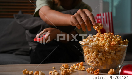 Shot of woman hand grabbing popcorn from a wooden bowl and watching TV at home. Leisure activity concept Shot of woman hand grabbing popcorn from a wooden bowl and watching TV at home. Leisure activity concept 92706184